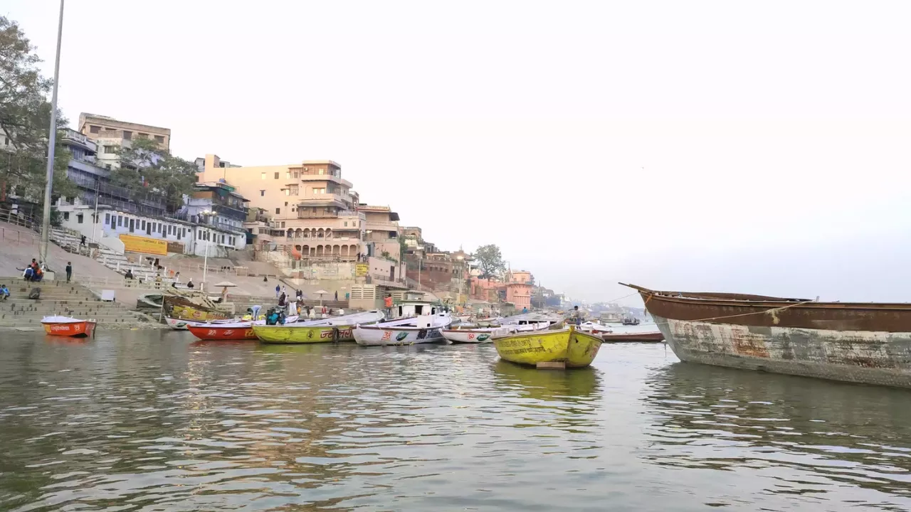 नदी पर लंगर डाले नौकाएँ; तट पर इमारतें और घाट, स्वच्छ आकाश के नीचे — वाराणसी: आत्मा की अन्तिम यात्रा की पावन भूमि